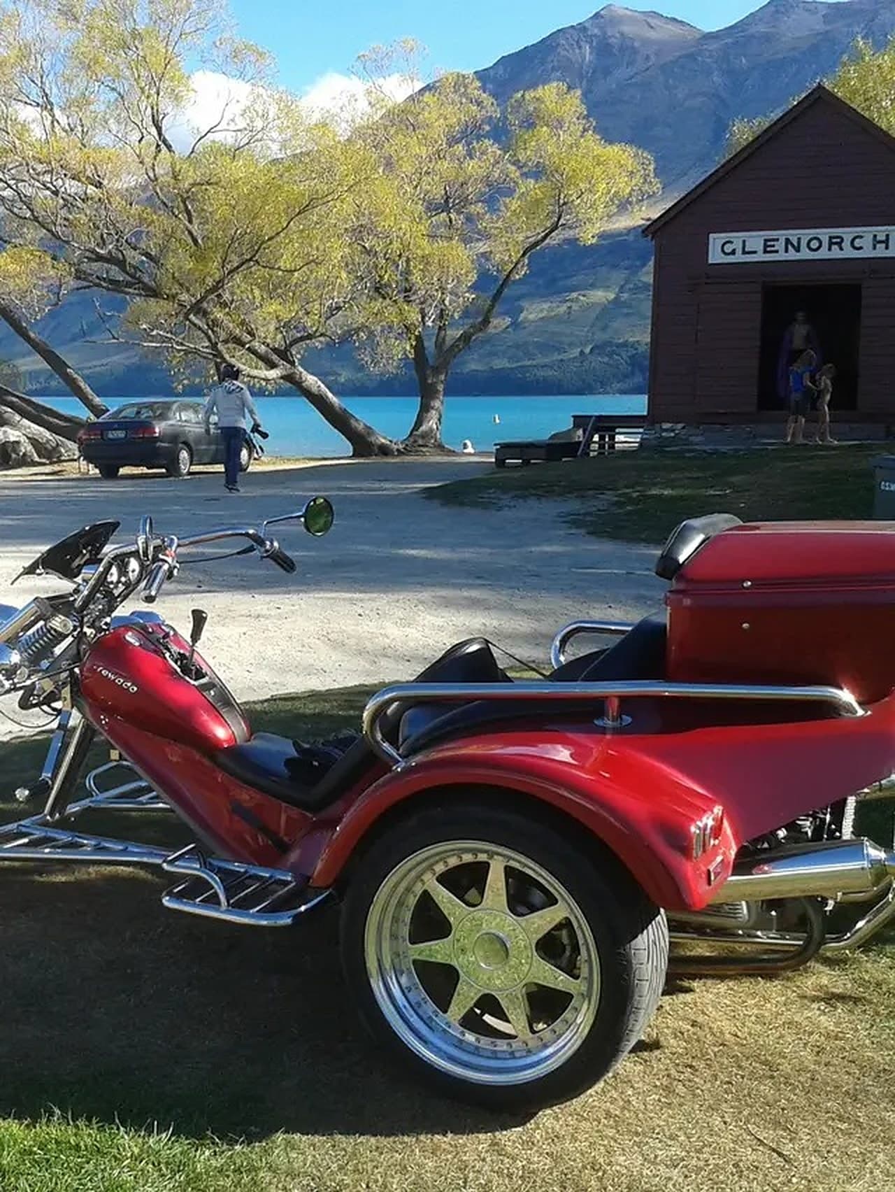 Red Rewaco trike parked beside Lake Wakatipu at Glenorchy, with autumn willows and mountains behind