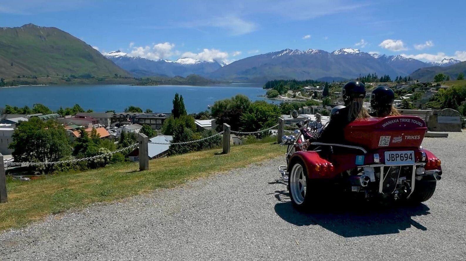 Red trike with two riders on a lakeside road, Wānaka town and mountains in the distance