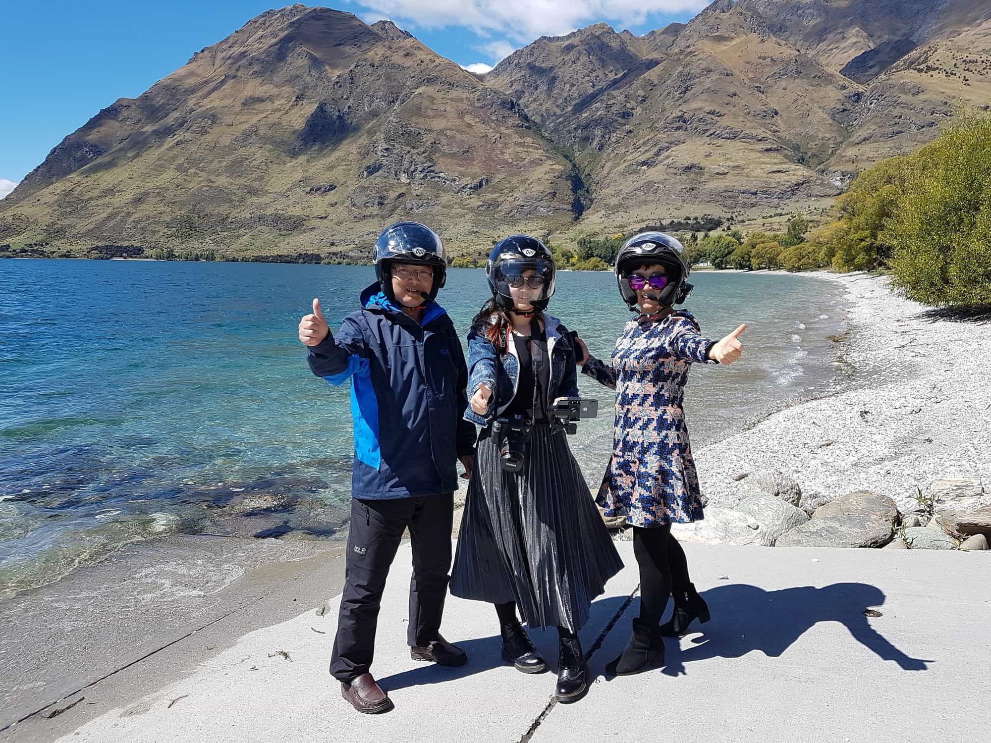 Three guests in helmets on a lake beach with mountains behind